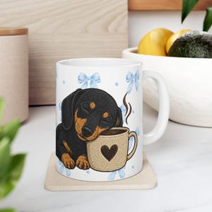 Könnte beinhalten: Weiße Keramik-Tasse mit einem schlafenden, schwarz-braunen Dackel-Welpen, der sich auf einer beigefarbenen Kaffeetasse mit einem Herz ausruht. Die Tasse hat blaue Schleifenakzente und Dampf, der aus der Tasse aufsteigt. Ein tolles Geschenk für Hundeliebhaber.