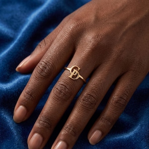 Peut inclure: Une bague dorée présentant deux lettres majuscules entrelacées, G et D, positionnées verticalement sur un anneau fin et lisse. Le métal possède une finition polie qui reflète la lumière, créant un design épuré et simple pour le doigt.