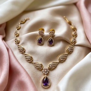 May include: A gold-toned necklace and matching earrings set. The necklace features a series of wing-shaped designs with clear crystals and a central teardrop-shaped purple gemstone. The earrings have a similar design with a purple teardrop gem.