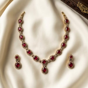May include: A gold-toned necklace and matching pair of earrings featuring oval-shaped red stones. Each stone is surrounded by a border of small clear crystals. The necklace has a central drop pendant, and the earrings consist of two stacked stones.