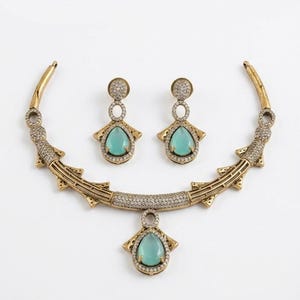 May include: A gold-toned necklace and matching earring set featuring teardrop-shaped light green stones. The jewelry is encrusted with small clear crystals and displays a textured, metallic finish with geometric accents along the collar.