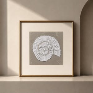 Puede incluir: Obra de arte enmarcada con un diseño de fósil de amonita blanco texturizado sobre un fondo gris. La obra está dentro de un marco dorado con un paspartú blanco, creando una estética sofisticada. Se exhibe sobre una repisa de tonos neutros.