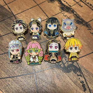 May include: A collection of eight gold-toned metal enamel pins featuring stylized anime characters. The pins depict various figures with distinct hair colors, facial expressions, and outfits, including a boar-headed character and a character with a bamboo muzzle.