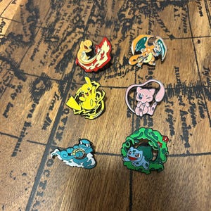 May include: A set of six colourful enamel pins featuring various animated creatures. The collection includes a yellow electric mouse, a pink feline, a blue turtle, a green plant-based creature, a fire-breathing dragon, and a small orange fire lizard.