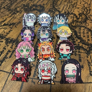 May include: A collection of eleven enamel pins featuring stylised anime characters with large eyes and colourful hair. Each pin depicts a different character with distinct facial features, hair colours, and accessories, arranged in a grid-like formation.