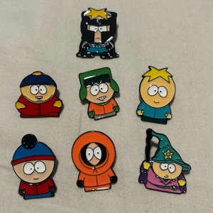 May include: Seven enamel pins featuring characters from the animated series South Park. The pins depict characters in various outfits, including a superhero, a wizard, and the main characters in their signature attire. The pins are colorful and detailed.