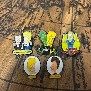 May include: Five enamel pins featuring Beavis and Butt-Head characters. One shows both characters with text, one shows them in green and yellow with Pink Floyd text, one shows Butt-Head with text Are You Threatening Me, and two individual portraits.