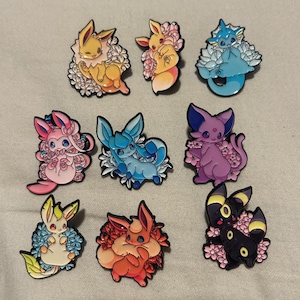 May include: A collection of nine colorful enamel pins featuring various cartoon characters. Each pin showcases a different character design, surrounded by floral accents. The pins are in shades of yellow, pink, blue, purple, orange, and black.