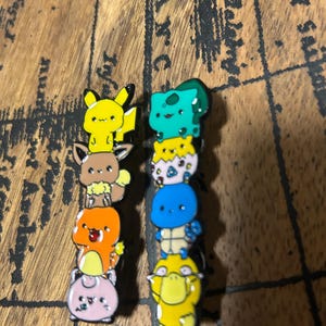 May include: Two vertical enamel pins featuring stacked cartoon characters. The left pin shows Pikachu, Eevee, Charmander, and Jigglypuff. The right pin displays Bulbasaur, Togepi, Squirtle, and Psyduck. Each character is rendered in vibrant colors.