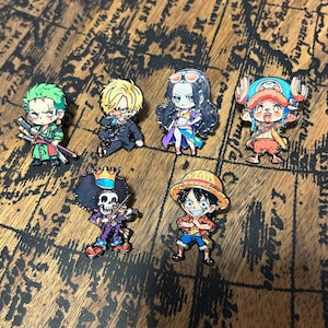 May include: A set of six enamel pins featuring chibi-style characters from the One Piece series, including Zoro with swords, Sanji, Robin, Chopper with blue antlers, Brook with a crown and violin, and Luffy wearing a straw hat and red shirt.