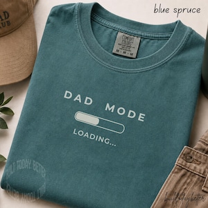 Puede incluir: Camiseta de color azul abeto con las palabras blancas "DAD MODE LOADING...". La camiseta tiene cuello redondo y una etiqueta que dice "COMFORT COLORS". La camiseta está doblada, junto a una gorra de béisbol beige y pantalones marrones.