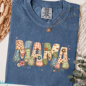 Puede incluir: Una camiseta azul Comfort Colors con la palabra "MAMA" en un diseño de patchwork. Las letras están hechas de varias telas florales y estampadas. Un collar dorado con un colgante descansa sobre la camiseta.