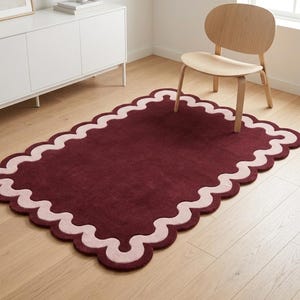 May include: A rectangular area rug featuring a deep burgundy center and a wide, light pink scalloped border. The rug has a tufted texture and is positioned on a light-colored wooden floor, showcasing a distinct wavy edge design throughout.