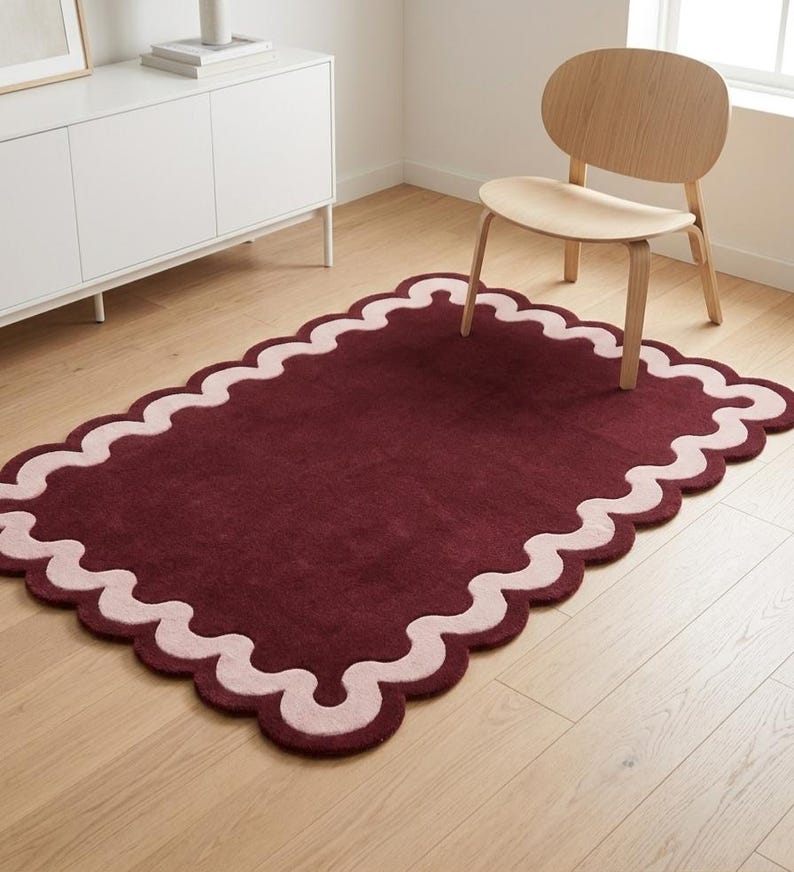May include: A rectangular area rug featuring a deep burgundy center and a wide, light pink scalloped border. The rug has a tufted texture and is positioned on a light-colored wooden floor, showcasing a distinct wavy edge design throughout.