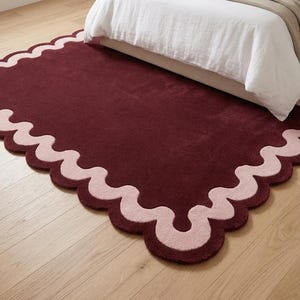 May include: A rectangular area rug featuring a deep burgundy center and a contrasting light pink scalloped border. The rug has a soft, plush texture and is positioned on a light-colored wooden floor, partially extending underneath a bed frame.