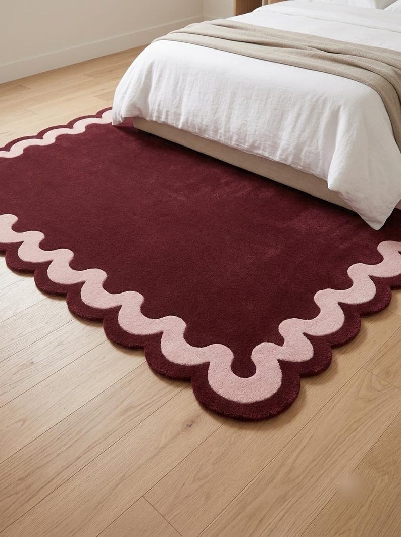 May include: A rectangular area rug featuring a deep burgundy center and a contrasting light pink scalloped border. The rug has a soft, plush texture and is positioned on a light-colored wooden floor, partially extending underneath a bed frame.