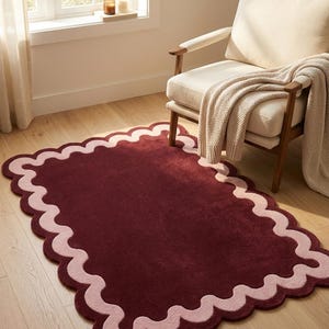 May include: A rectangular area rug featuring a deep burgundy center and a wide, scalloped border in a soft light pink hue. The rug has a plush, tufted texture and is positioned on a light-toned wooden floor, showcasing a distinct wavy edge design.