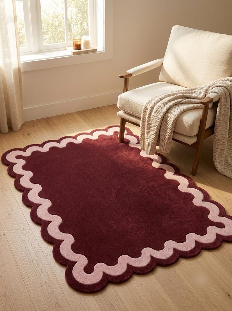 May include: A rectangular area rug featuring a deep burgundy center and a wide, scalloped border in a soft light pink hue. The rug has a plush, tufted texture and is positioned on a light-toned wooden floor, showcasing a distinct wavy edge design.