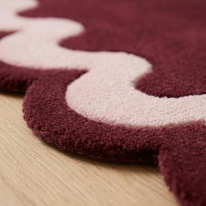 May include: A plush area rug featuring a deep burgundy base color with a contrasting light pink wavy border. The rug has a thick, tufted texture and a scalloped edge design that follows the curved lines of the light pink pattern throughout.