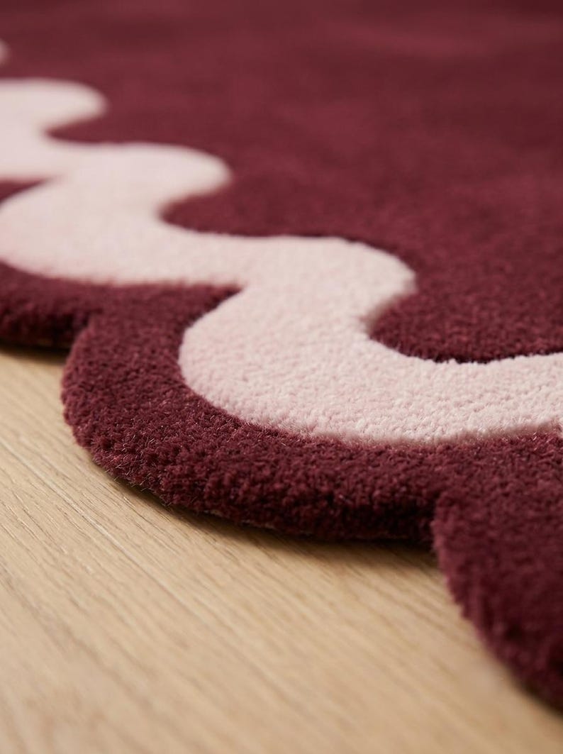 May include: A plush area rug featuring a deep burgundy base color with a contrasting light pink wavy border. The rug has a thick, tufted texture and a scalloped edge design that follows the curved lines of the light pink pattern throughout.