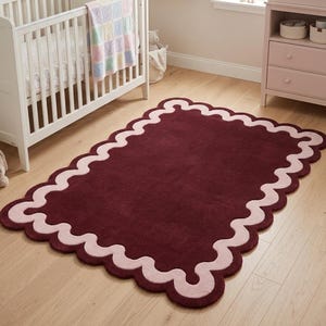 May include: A rectangular area rug featuring a deep burgundy center and a wide, scalloped border in a soft pastel pink. The rug has a plush, tufted texture and a continuous wavy edge design that frames the solid dark red interior surface.