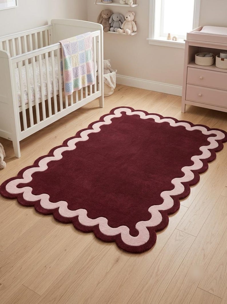 May include: A rectangular area rug featuring a deep burgundy center and a wide, scalloped border in a soft pastel pink. The rug has a plush, tufted texture and a continuous wavy edge design that frames the solid dark red interior surface.
