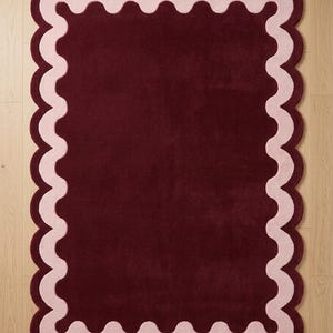 May include: A rectangular area rug featuring a deep burgundy center and a wide, light pink scalloped border. The rug has a soft, tufted texture and a consistent pile height. A small white square label in the corner displays the text Paradise Carpets.