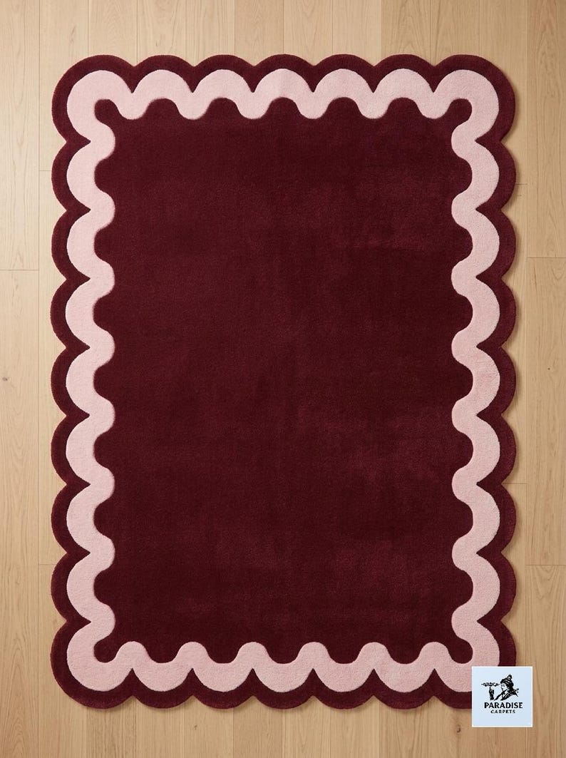 May include: A rectangular area rug featuring a deep burgundy center and a wide, light pink scalloped border. The rug has a soft, tufted texture and a consistent pile height. A small white square label in the corner displays the text Paradise Carpets.