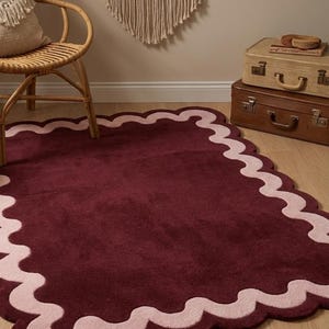 May include: A rectangular area rug featuring a deep burgundy center and a wide, scalloped border in a soft, light pink hue. The rug has a plush, tufted texture and is laid flat on a light-colored wooden floor, showcasing its distinct wavy edges.