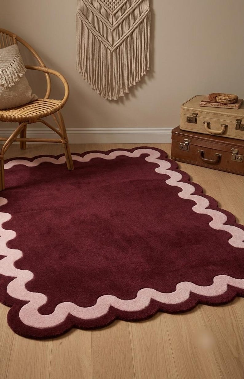 May include: A rectangular area rug featuring a deep burgundy center and a wide, scalloped border in a soft, light pink hue. The rug has a plush, tufted texture and is laid flat on a light-colored wooden floor, showcasing its distinct wavy edges.