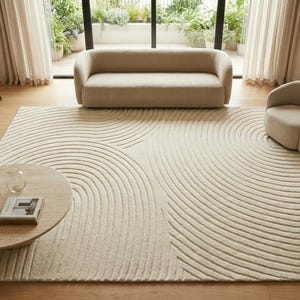 May include: A beige living room rug with a circular pattern dominates the space. A beige sofa and two armchairs complement the rug. A round wooden coffee table with books and a vase sits in the foreground. The room has large windows and beige curtains.