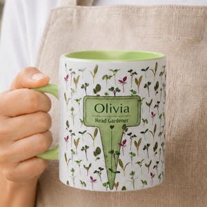May include: A white ceramic mug with a light green interior and handle, featuring a repeating pattern of small green and purple sprouts. A central green garden stake graphic displays the text Olivia and Head Gardener in black lettering.