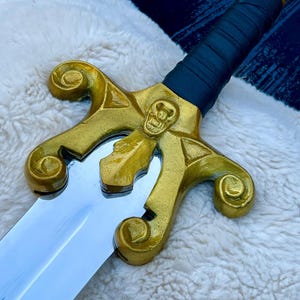May include: A decorative sword with a gold-coloured hilt featuring a skull design and a polished silver blade. The handle is wrapped in dark blue material. The sword rests on a white, fluffy surface.