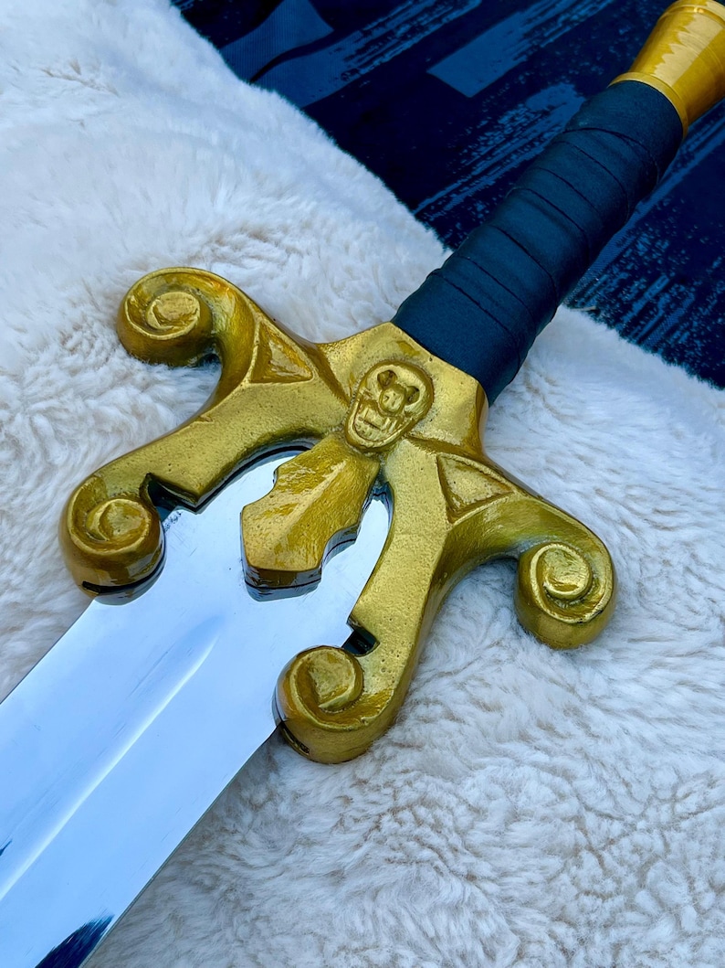 May include: A decorative sword with a gold-coloured hilt featuring a skull design and a polished silver blade. The handle is wrapped in dark blue material. The sword rests on a white, fluffy surface.