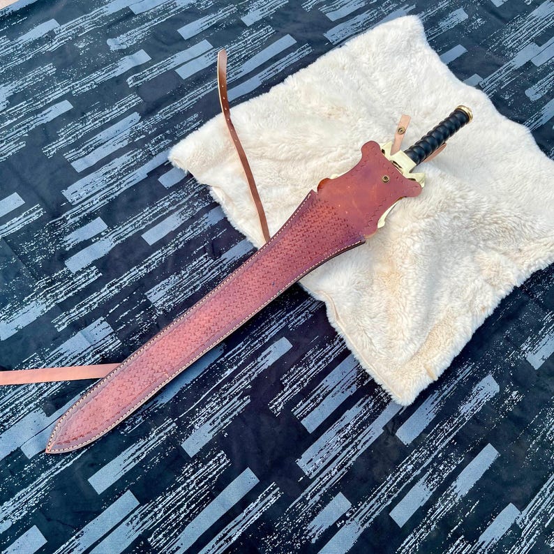 Pu&ograve; includere: Un lungo fodero per spada in pelle marrone con una spada abbinata. La spada ha un'impugnatura nera e dettagli dorati. Il fodero &egrave; appoggiato su una superficie bianca e soffice, con uno sfondo nero e grigio a motivi.