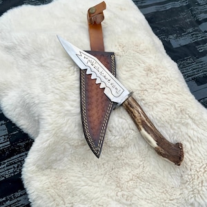 May include: A hunting knife with a serrated blade and a handle made of deer antler. The blade has etched text and is paired with a brown leather sheath. The sheath has a decorative pattern and a leather strap.