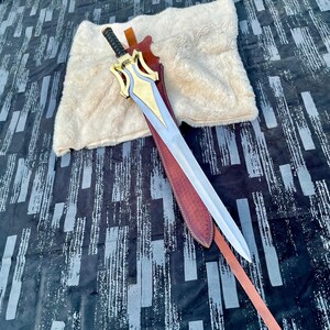 Pu&ograve; includere: Una spada argentata con un'elsa dorata e blu &egrave; in un fodero di cuoio marrone. La spada ha una lama lunga e dritta e un'impugnatura decorativa. Il fodero &egrave; rifinito con cuciture e un cinturino in pelle. Un panno soffice color crema &egrave; sullo sfondo.