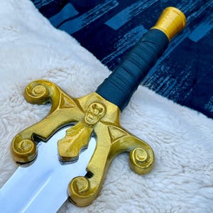May include: A decorative sword with a gold-coloured hilt featuring a skull design and a silver blade. The handle is wrapped in dark blue material, and the pommel is gold. The sword is displayed on a white, fluffy surface.