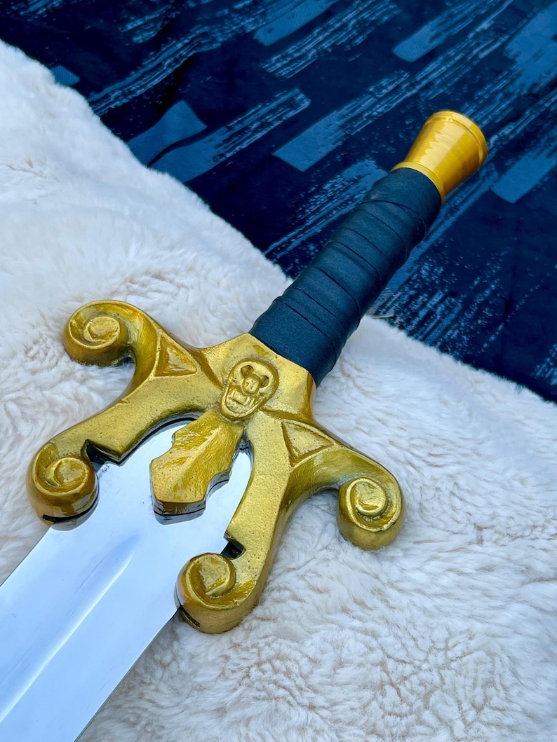 May include: A decorative sword with a gold-coloured hilt featuring a skull design and a silver blade. The handle is wrapped in dark blue material, and the pommel is gold. The sword is displayed on a white, fluffy surface.