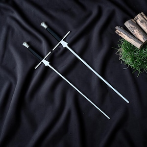 May include: Two long, slender metal skewers with black handles and silver-toned pommels. Each skewer features a crossguard design near the handle, resembling a miniature sword. The metal blades are straight, thin, and pointed at the ends.