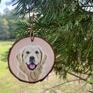 May include: A round wooden ornament featuring a painted portrait of a Golden Retriever dog. The dog has a light brown coat, a pink tongue, and is smiling. The ornament is hanging from a brown leather cord.