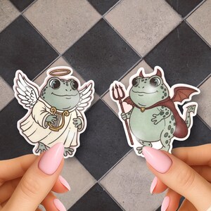 May include: Two die-cut stickers featuring green frogs. One frog is dressed as an angel with white wings, a halo, and a golden harp. The other frog is dressed as a devil with small red horns, red wings, a red cape, and a brown trident.