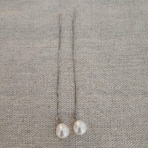Puede incluir: Un par de pendientes colgantes de plata con una perla blanca en forma de lágrima. Los pendientes cuelgan verticalmente, mostrando la belleza natural de las perlas. Una joya sencilla y elegante, ideal para cualquier ocasión.