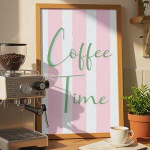 Könnte beinhalten: Ein gerahmter Druck mit den Worten "Coffee Time" in grüner Schrift auf einem rosa-weiß gestreiften Hintergrund. Eine Kaffeemaschine, eine Kaffeetasse und ein offenes Buch befinden sich auf einer Holztheke.