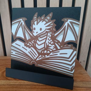 Peut inclure: Un présentoir de livre noir avec un motif de dragon marron et blanc. Le dragon est assis sur un livre ouvert. Le présentoir est en métal et est posé sur une surface en bois.