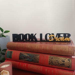 Peut inclure: Trois livres anciens aux couvertures rouges et dorées sont empilés. Un signe noir "BOOK LOVER" avec l'inscription dorée "dream" est placé au-dessus. Une bougie allumée dans un verre rose est visible à gauche.