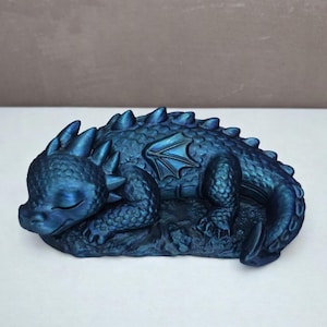Pode incluir: Estatueta de dragão azul adormecido com reflexos iridescentes. O dragão está enroscado, com os olhos fechados e as asas visíveis. A estatueta está sobre uma base texturizada, mostrando escamas e detalhes.