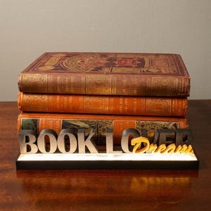 Może przedstawiać: Stos dwóch antycznych książek z ozdobnymi okładkami, zwieńczony trzecią książką. Z przodu dekoracyjny napis "BOOK LOVER Dream" w srebrnych i złotych literach, na czarnej podstawie.