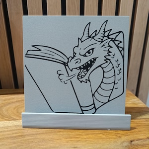 Peut inclure: Plaque carrée grise avec un dessin au trait noir d'un dragon lisant un livre. Le dragon est orienté vers la gauche, la bouche ouverte et de la fumée s'échappant. La plaque est dans un support argenté.