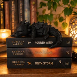 Può includere: Una statuina di drago nero è appoggiata su una pila di tre libri. I libri sono intitolati "Fourth Wing", "Iron Flame" e "Onyx Storm" di Rebecca Yarros. Il drago ha le ali.